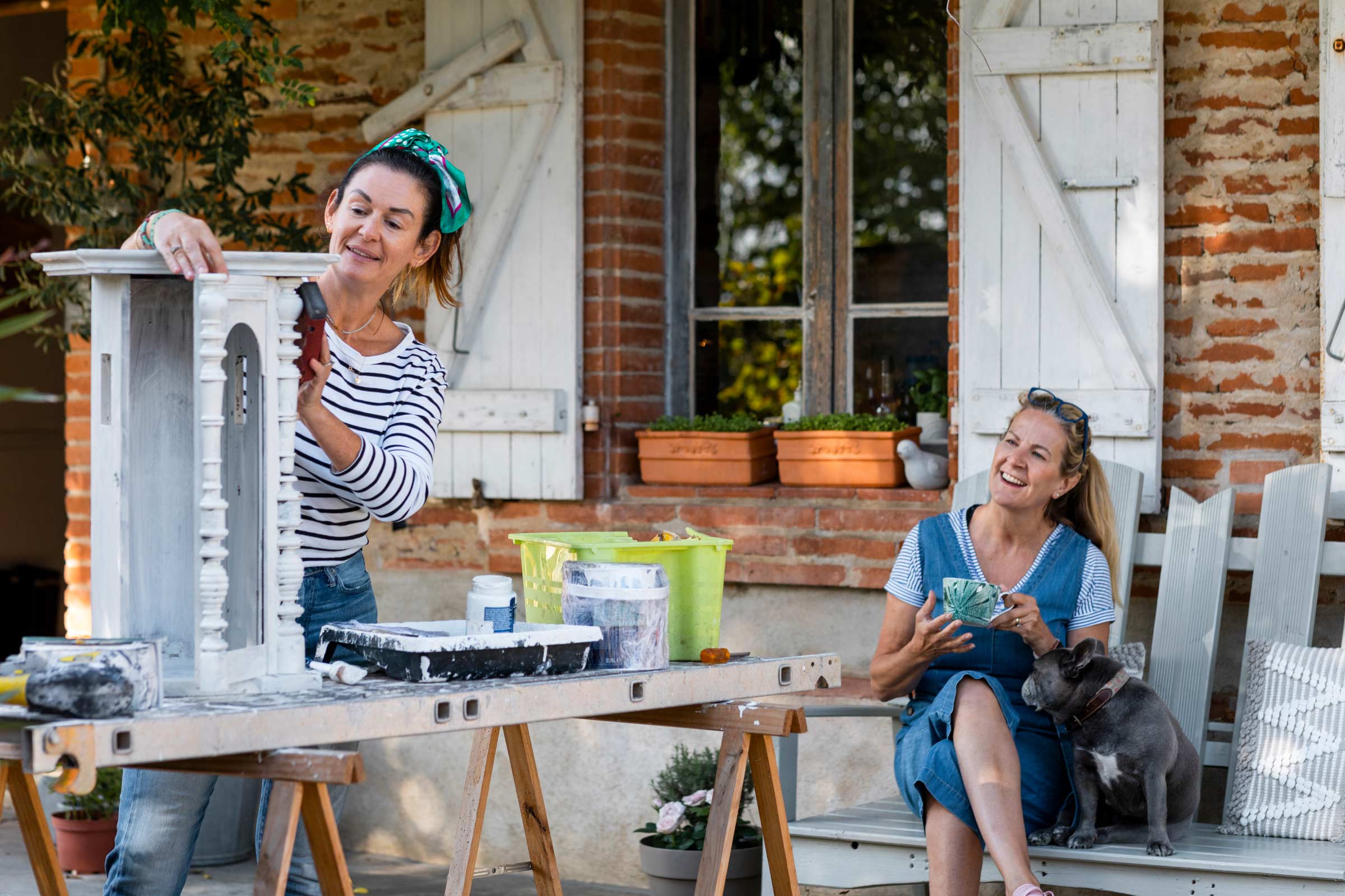 Two Caucasian women outside working on DIY patio projects by sanding and painting furniture.